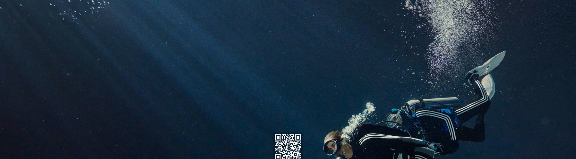 A scuba diver wearing a black wetsuit with white adidas stripes swims near colourful coral.