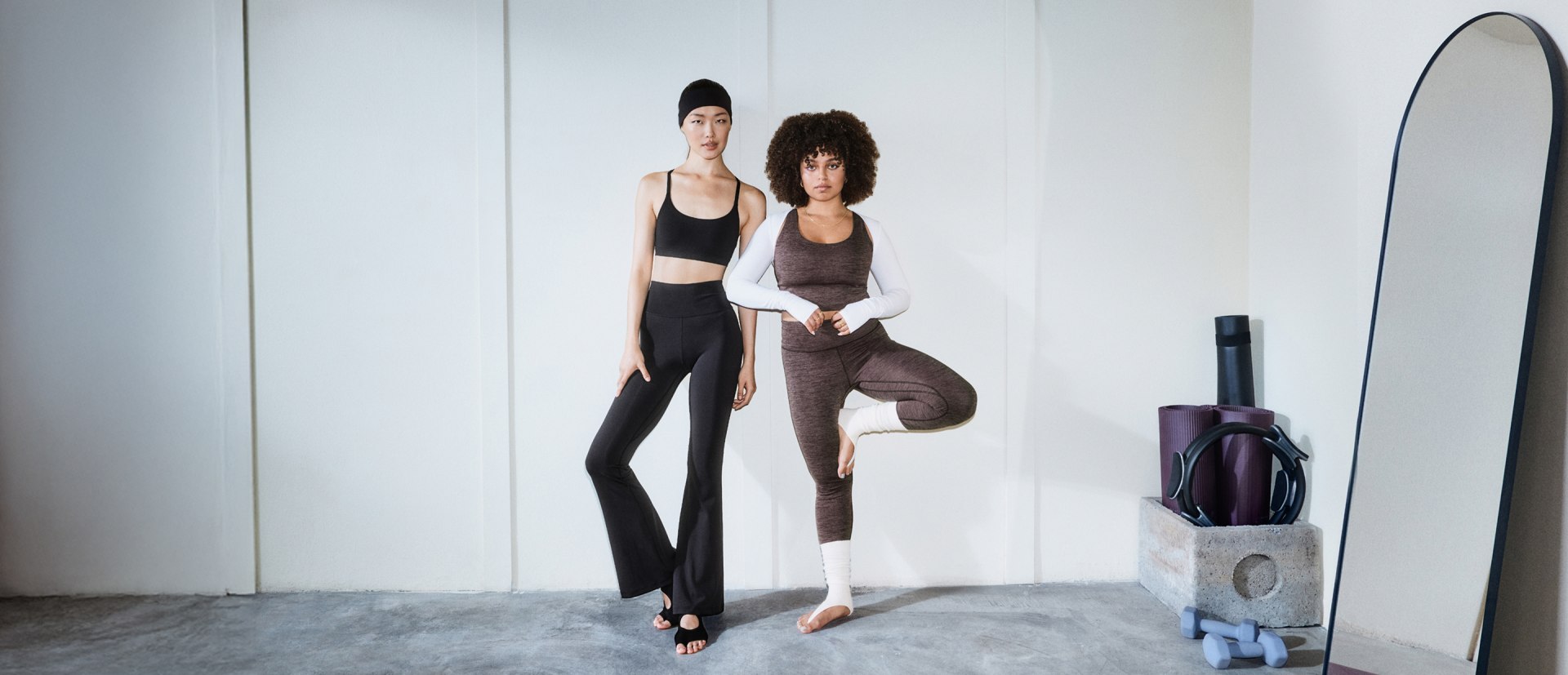 Due donne in abbigliamento sportivo in piedi, in una stanza minimalista con pareti bianche e un pallone da allenamento viola sullo sfondo.