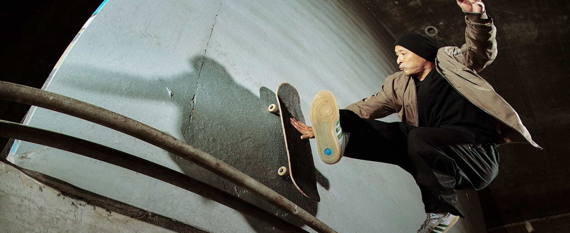 A skateboarder wearing white and green adidas Glenburn sneakers, black pants, a black beanie, and a light brown jacket performs a wall ride trick in an urban skate spot.