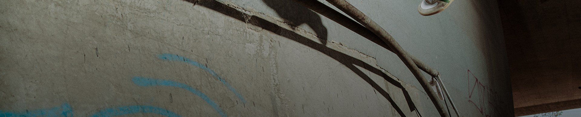 A skateboarder wearing white and green adidas Glenburn sneakers, black pants, a black beanie, and a light brown jacket performs a wall ride trick in an urban skate spot.