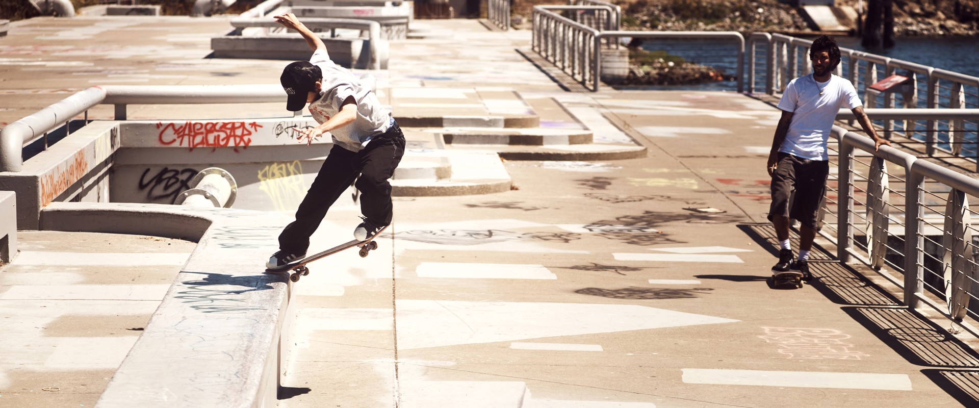 A skateboarder in a white t-shirt and black pants stands by a waterfront rail, wearing adidas Campus ADV trainers, shown up close next to a worn skateboard.