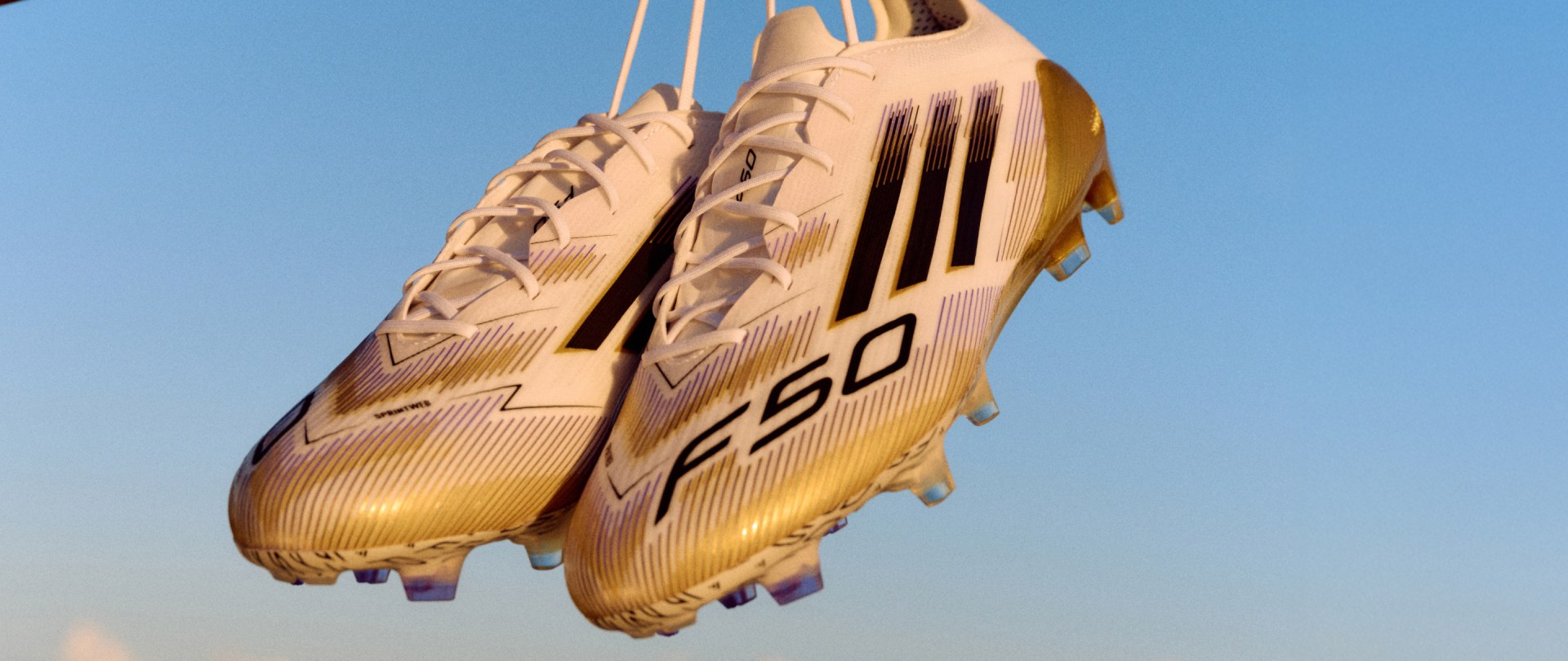 A pair of gold-colored soccer cleats with distinctive patterns and the "F50" logo are suspended against a clear blue sky.
