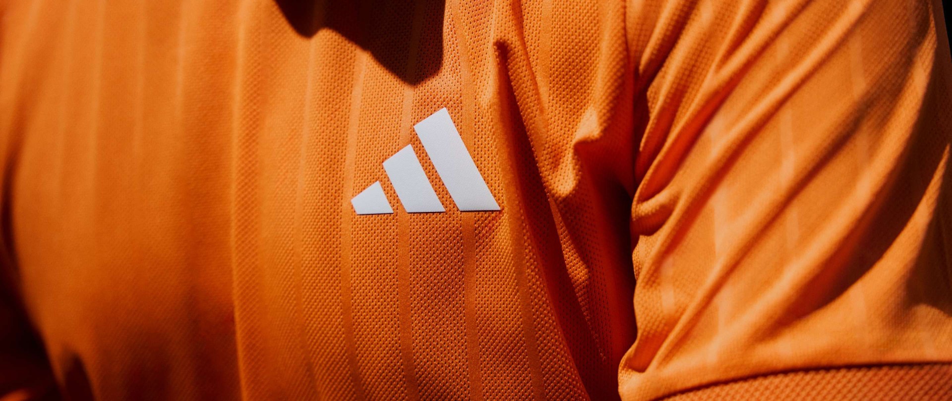 La imagen muestra una camiseta deportiva naranja con el logotipo de adidas en contraste.