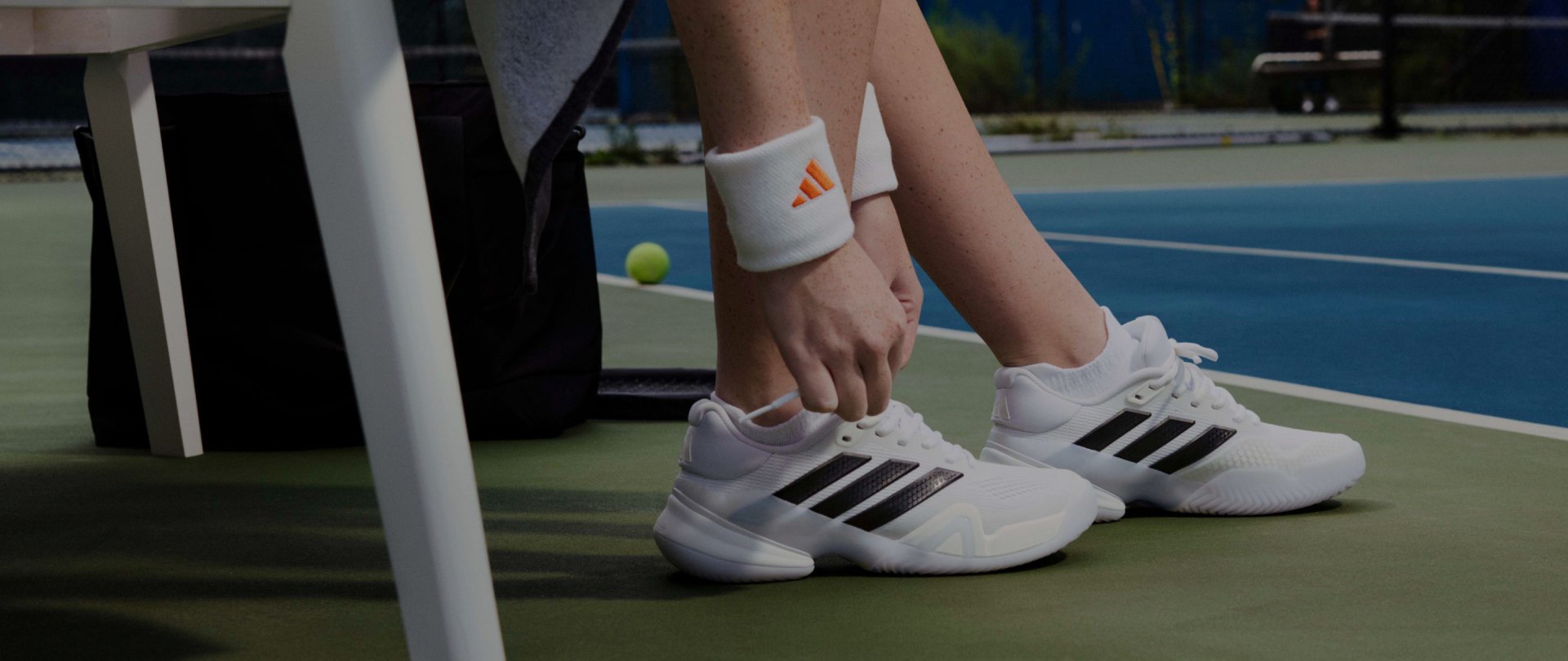 ภาพนี้แสดงให้เห็นถึงขาและเท้าของคนสวมรองเท้าเทนนิสสีขาวที่มีลายแถบสีดำบนสนามเทนนิสสีน้ำเงิน