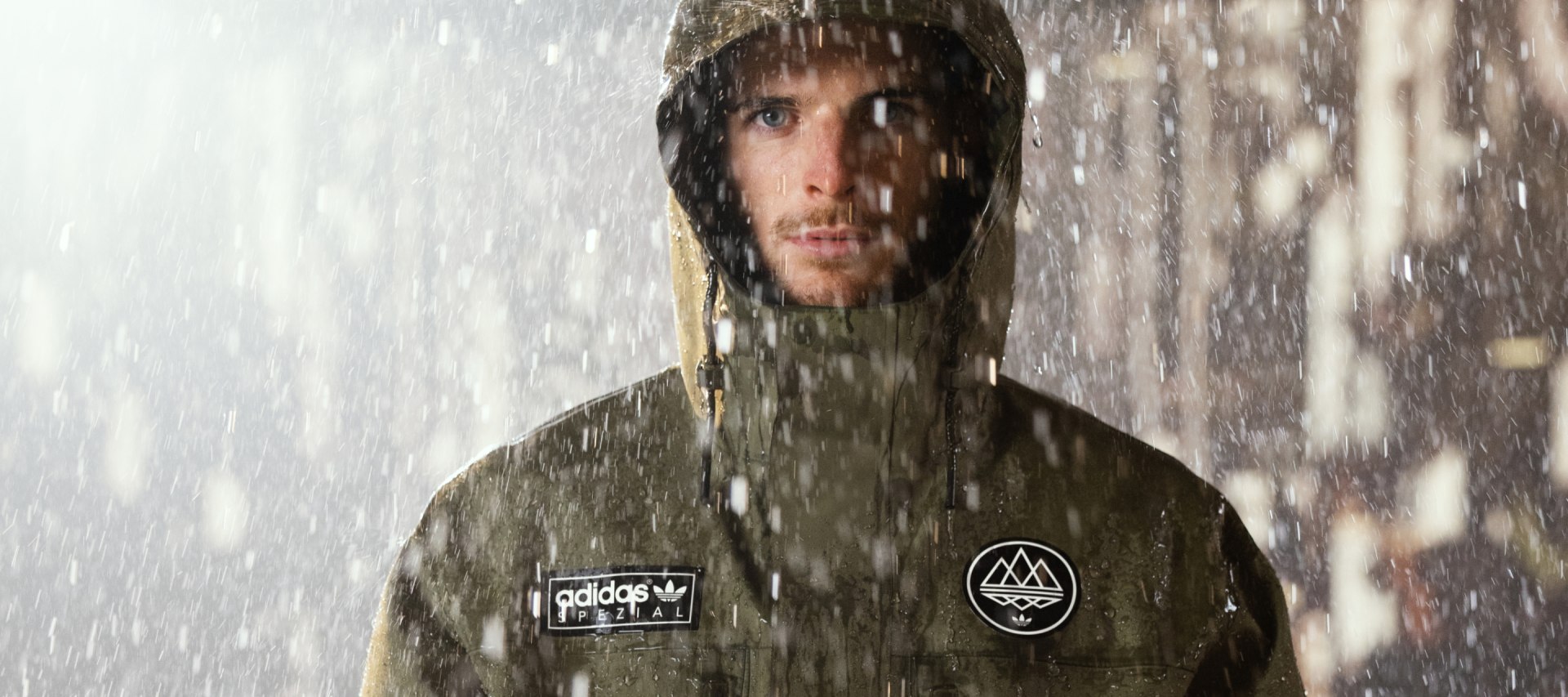 A person wearing a hooded jacket stands in the rain, with water droplets visible on the jacket and face.