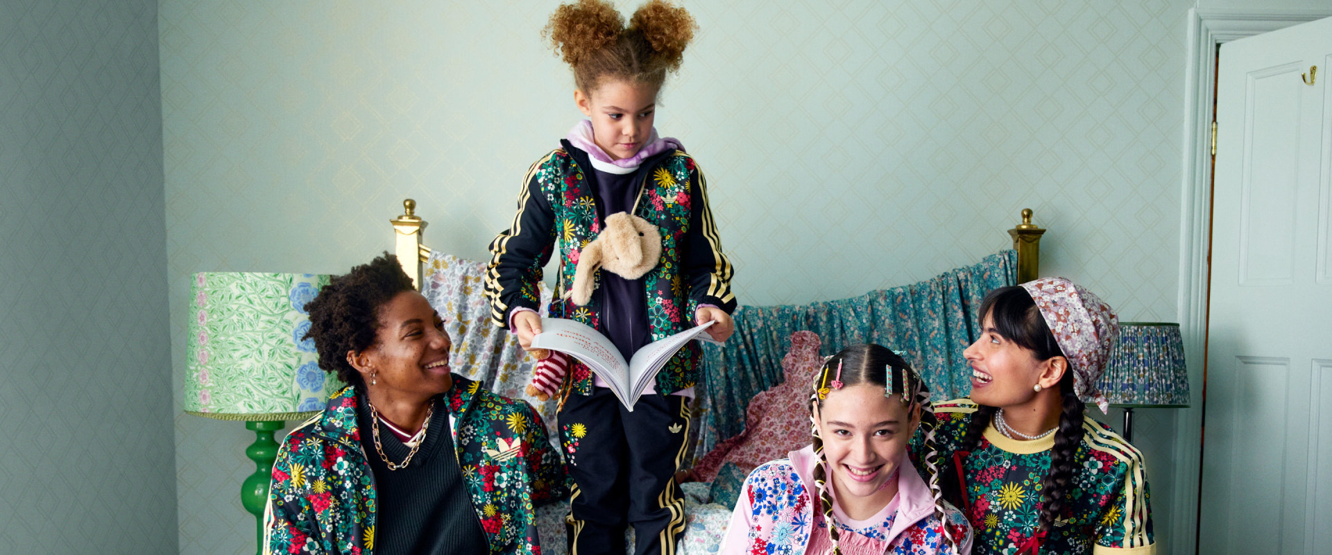 A family is sitting on a bed in their family home, they are smiling at each other and the youngest daughter is holding a book. Both mothers and their two daughters are wearing adidas x Liberty London.