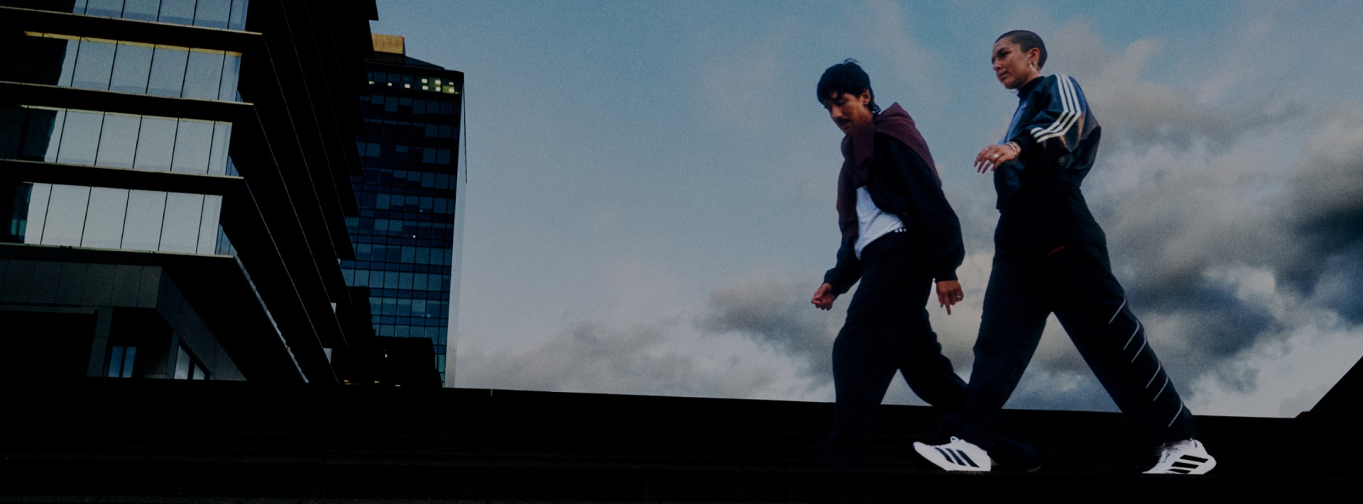 Two individuals in dark clothing stand on a rooftop overlooking a cityscape with tall buildings in the background.