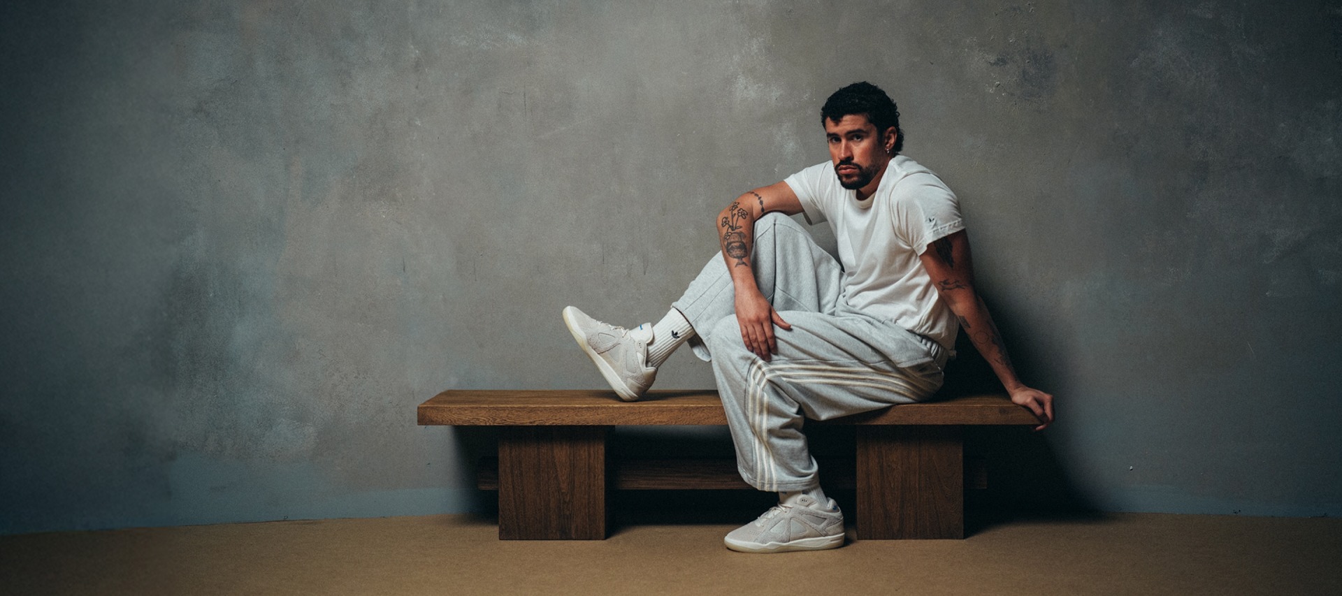 Bad Bunny mira a la cámara, sentado en un banco de madera contra una pared gris texturizada, usando una camiseta blanca, pantalones deportivos adidas gris claro, medias adidas blancas y zapatillas adidas blancas. Una pierna está doblada en la banca y el otro pie descansa en el suelo.