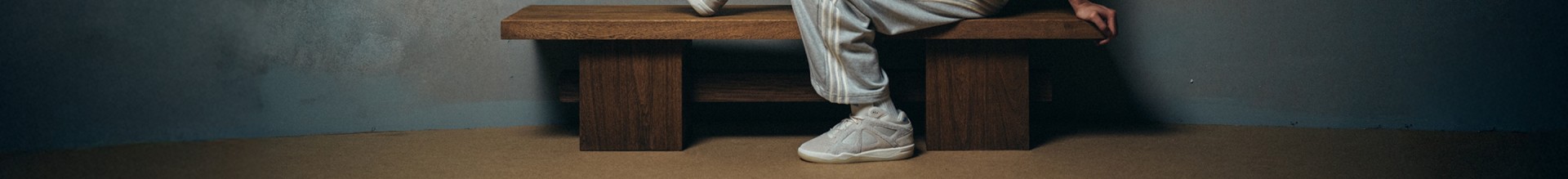 Bad Bunny looks at the camera, sitting on a wooden bench against a textured gray wall, wearing a white T-shirt, light gray adidas track pants, white adidas socks, and white adidas sneakers. One leg is bent on the bench, the other foot rests on the floor.