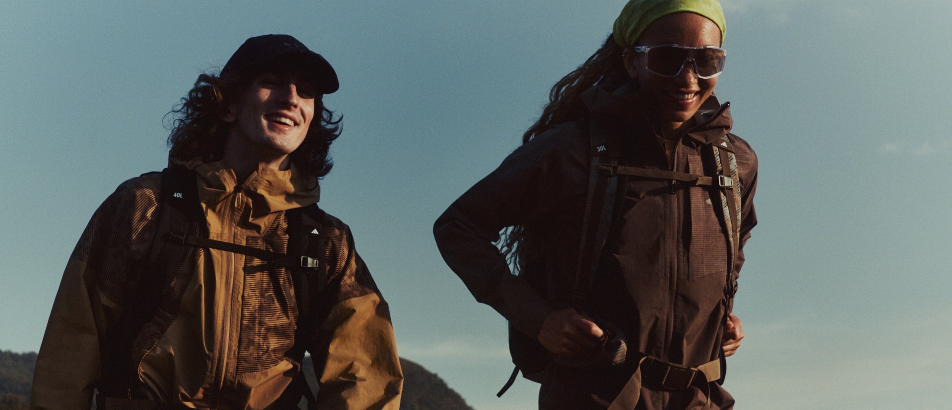 Zwei Personen, die dunkle Kapuzenjacken und Sonnenbrillen tragen, stehen vor einem strahlend blauen Himmel.