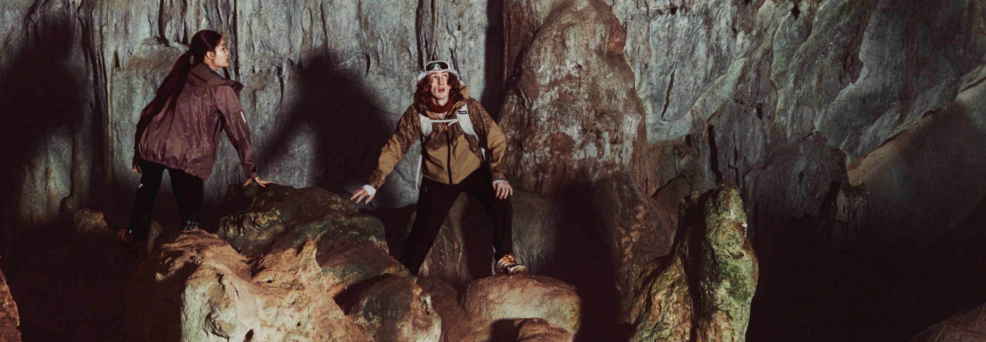 L'image montre deux individus debout dans un environnement sombre et rocheux semblable à une grotte, avec en arrière-plan des formations rocheuses complexes et texturées.