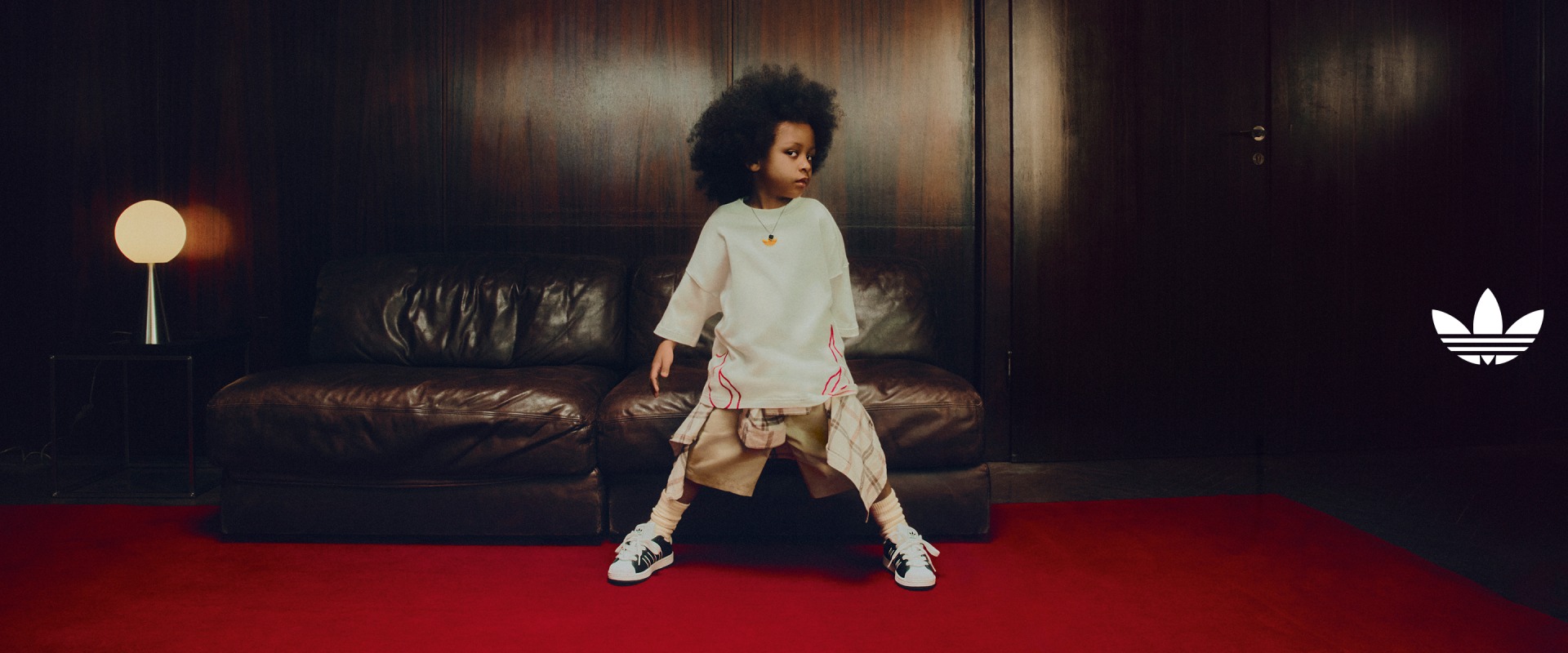 A child wearing black-and-white adidas Superstar sneakers stands confidently on a red carpet in front of a leather sofa, with the Trefoil logo overlayed to the right side.