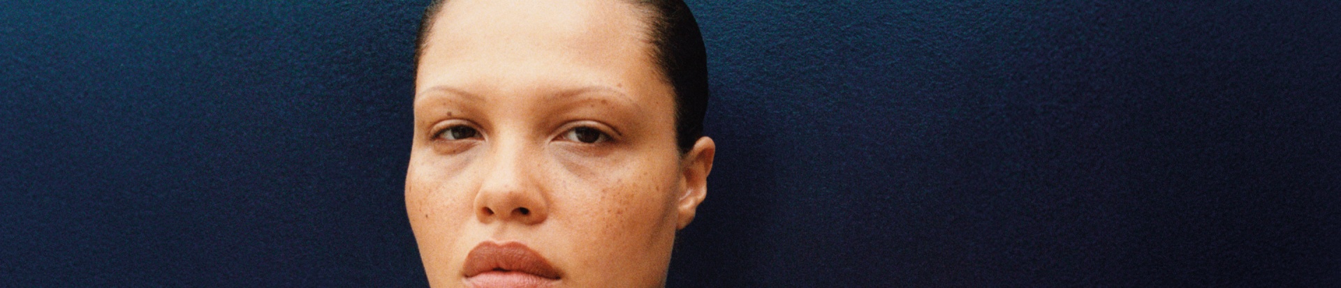 A person wearing a blue adidas x Wales Bonner zip-up jacket with white stripes and the Trefoil logo on the chest, standing against a dark blue background.