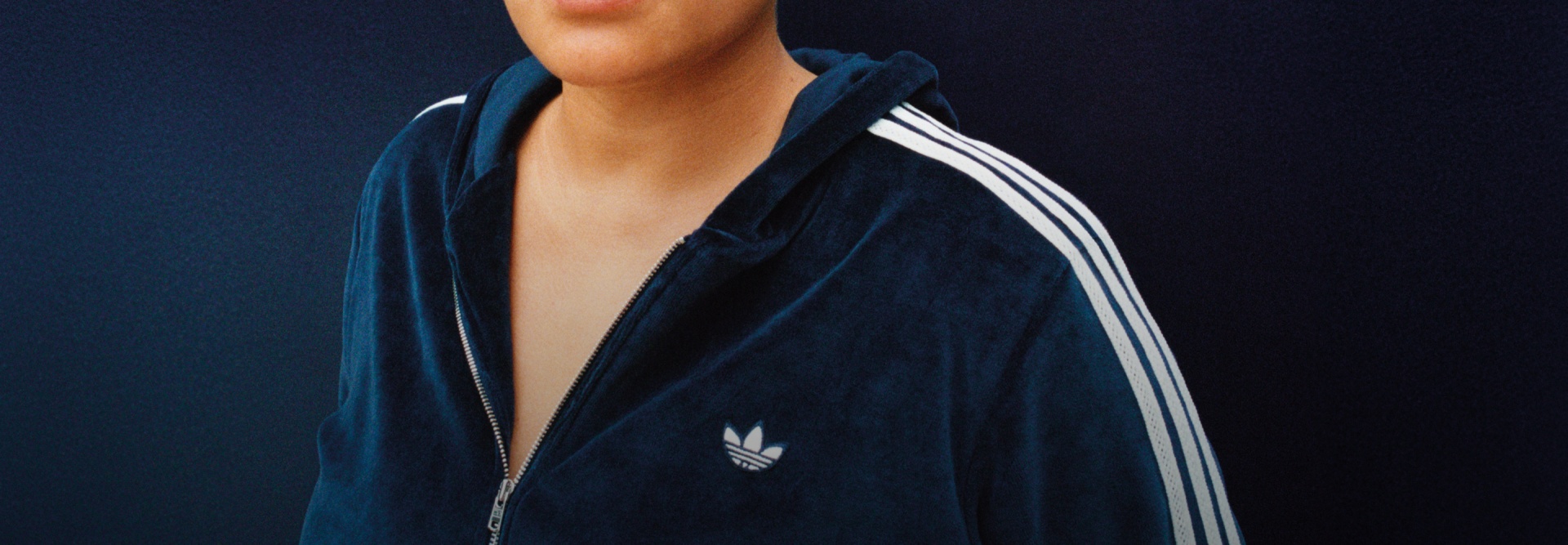 A person wearing a blue adidas x Wales Bonner zip-up jacket with white stripes and the Trefoil logo on the chest, standing against a dark blue background.
