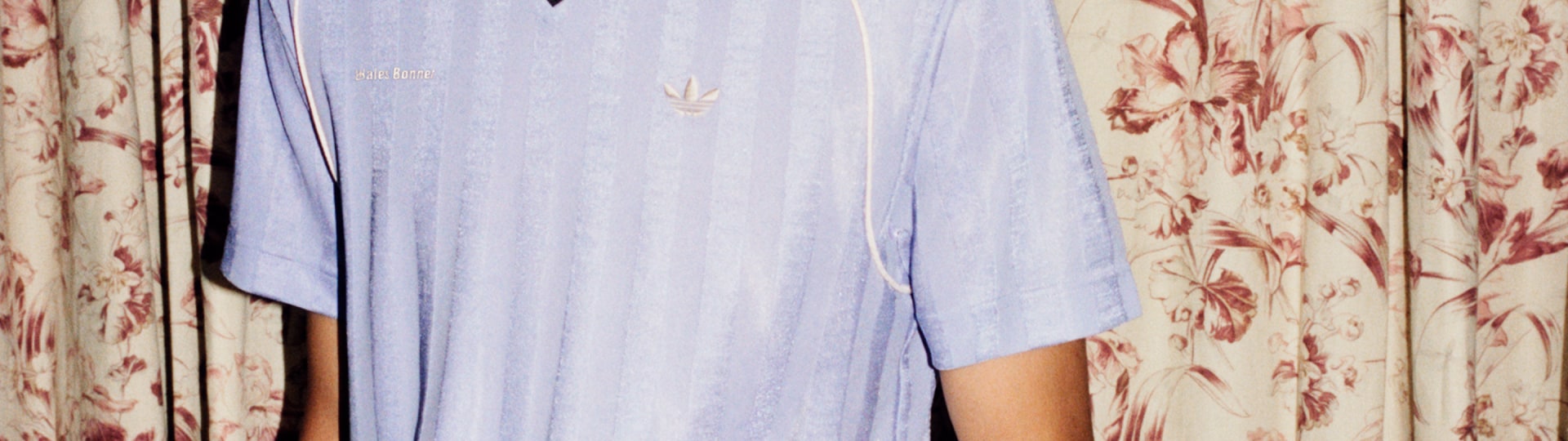 A person wearing a light blue adidas x Wales Bonner jersey with subtle vertical stripes, a black collar, and the Trefoil logo on the chest, standing in front of floral-patterned curtains.