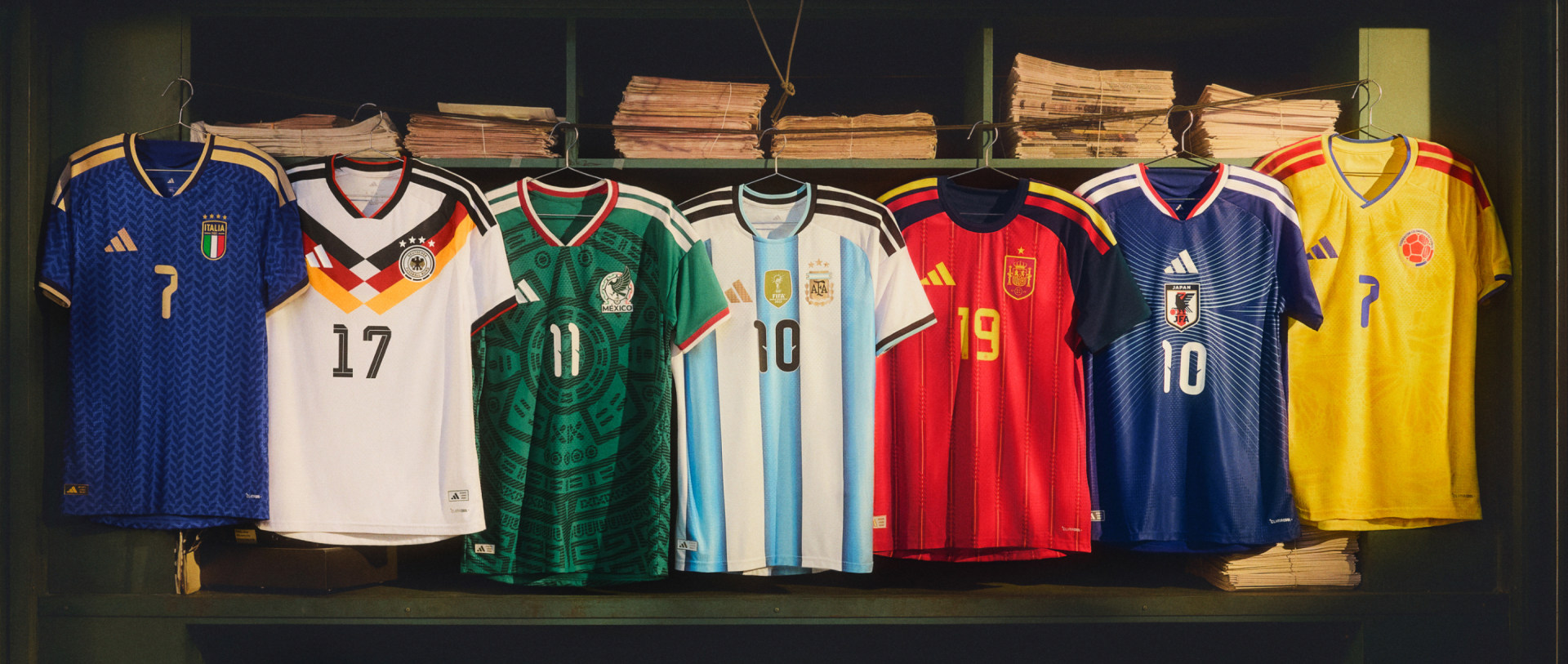A collection of colorful soccer jerseys from various national teams, displayed on a wooden shelf against a dark background with stacks of papers or documents in the background.