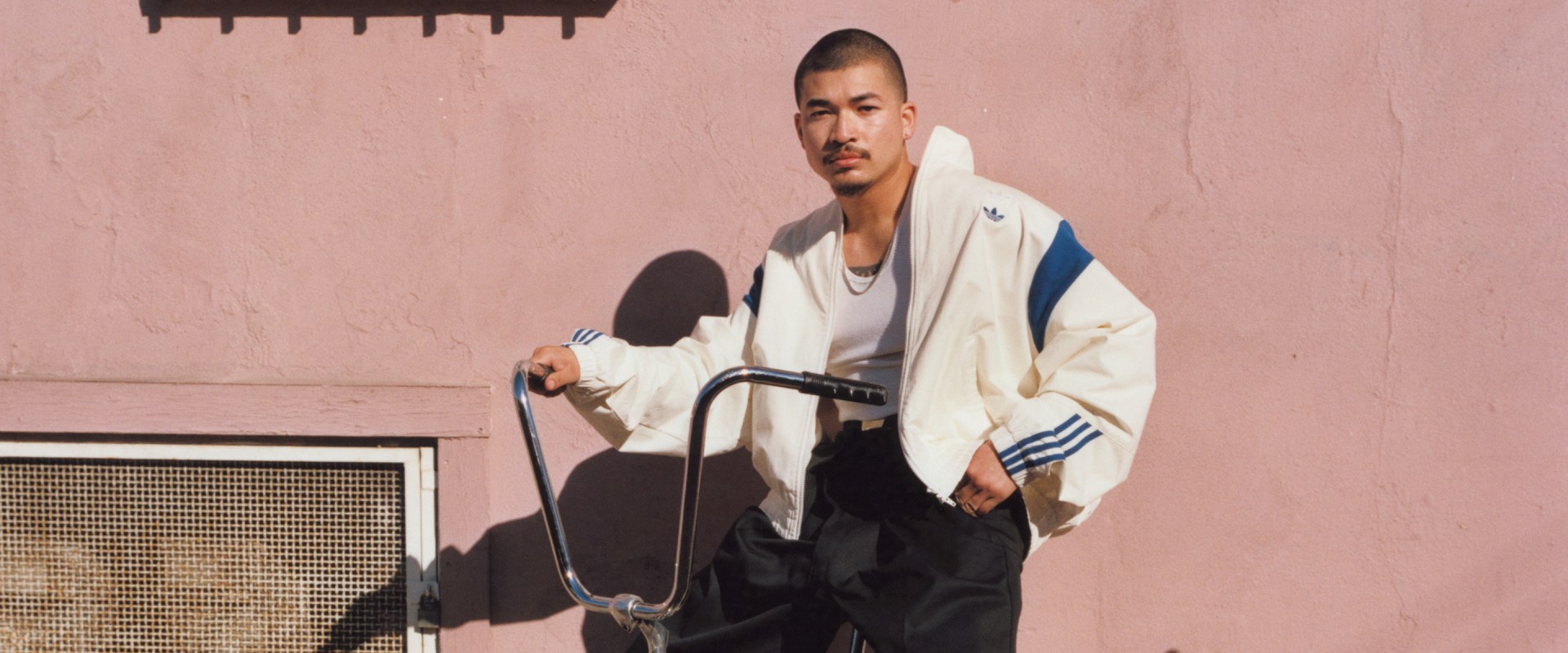 A man stands beside a bicycle in front of a pink wall, wearing a cream adidas x Willy Chavarria jacket with blue accents and black pants.