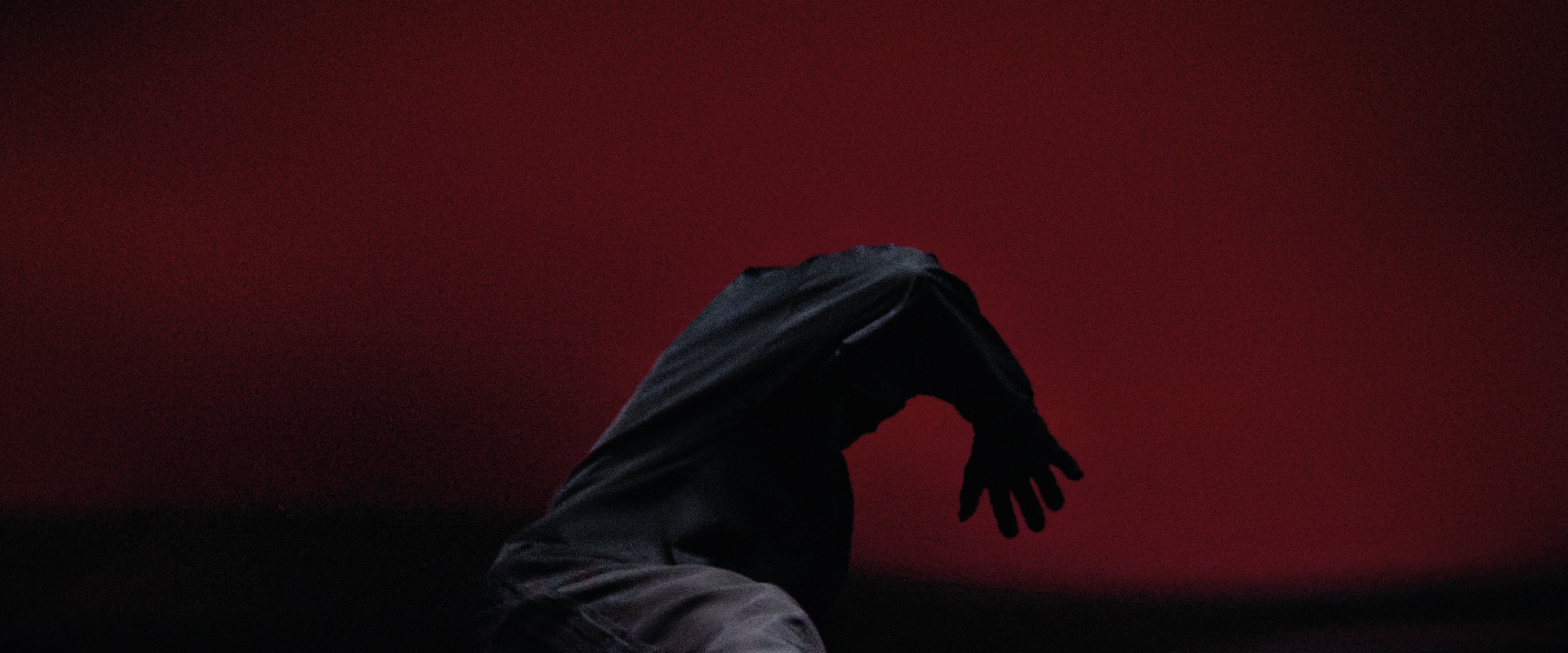 En mørk, skyggefull figur med utstrakt hånd kommer fra en dyp, rød bakgrunn.