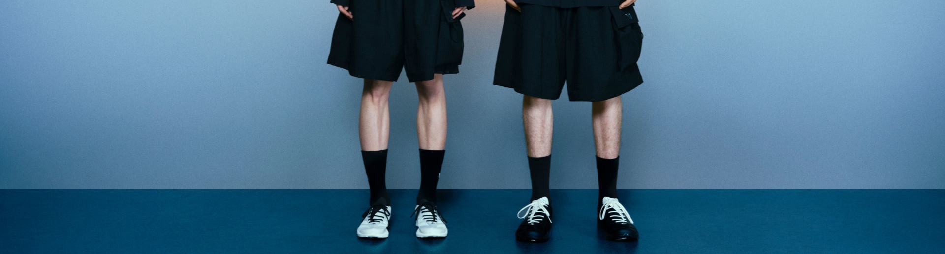 The image features two pairs of legs wearing black shorts and black socks, with one pair wearing white trainers and the other pair wearing black dress shoes, set against a plain blue background.