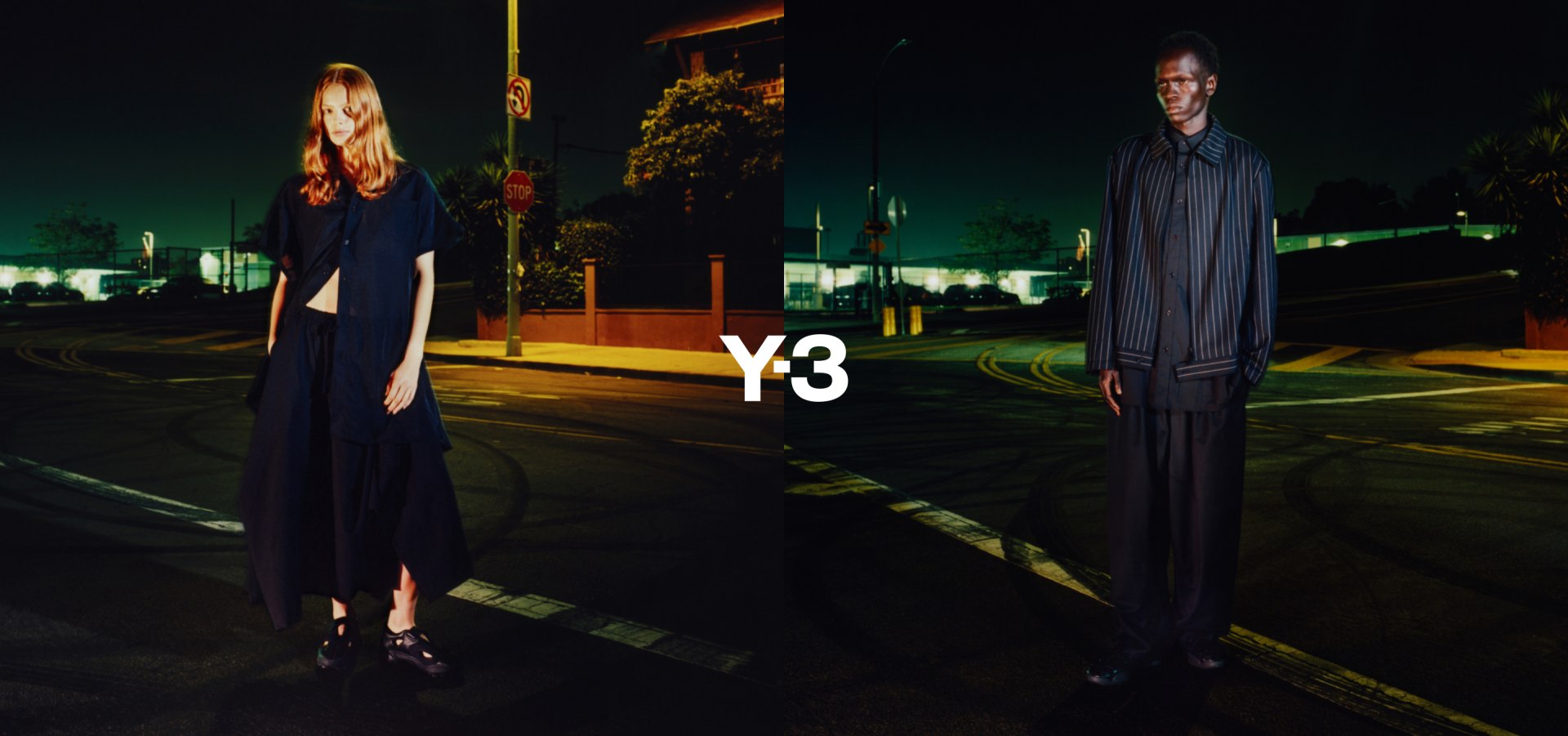 Two individuals, a woman in a long dark dress and a man in a long dark coat, stand in a dimly lit outdoor setting with a large "Y3" sign visible in the background.