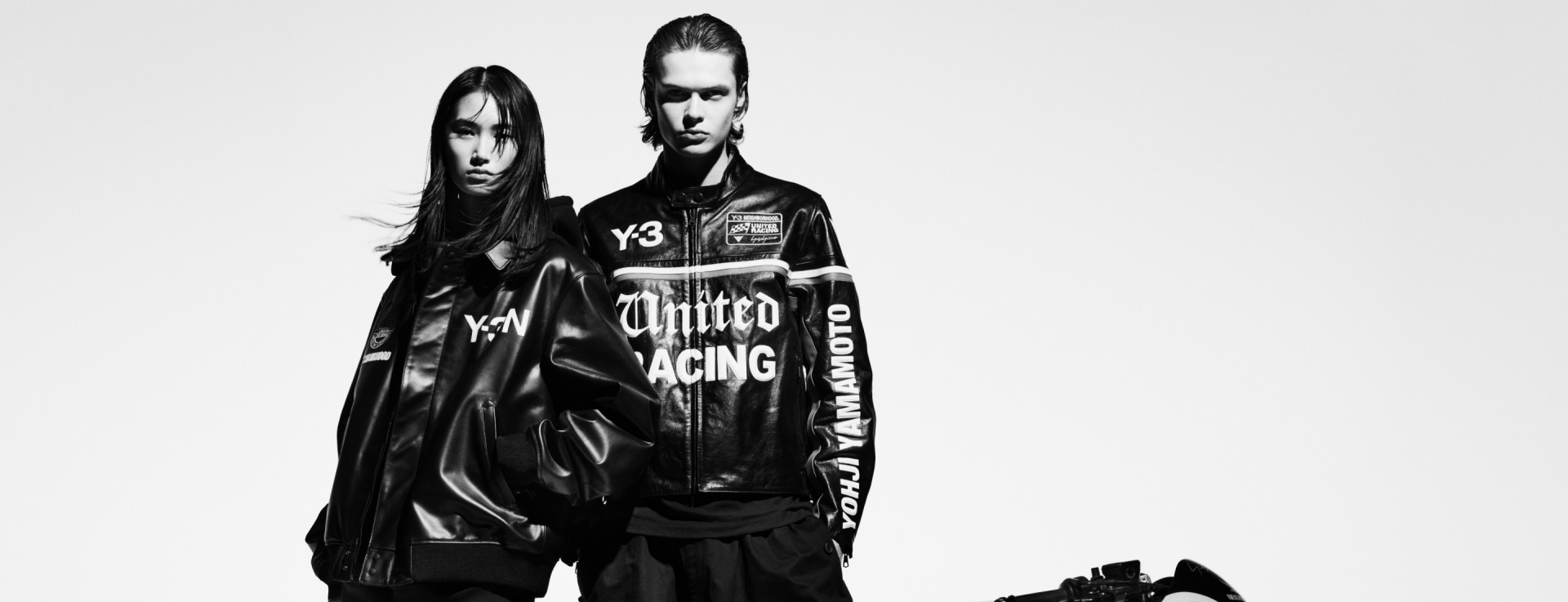 Dos individuos vestidos de negro se paran frente a una motocicleta en una imagen en blanco y negro.