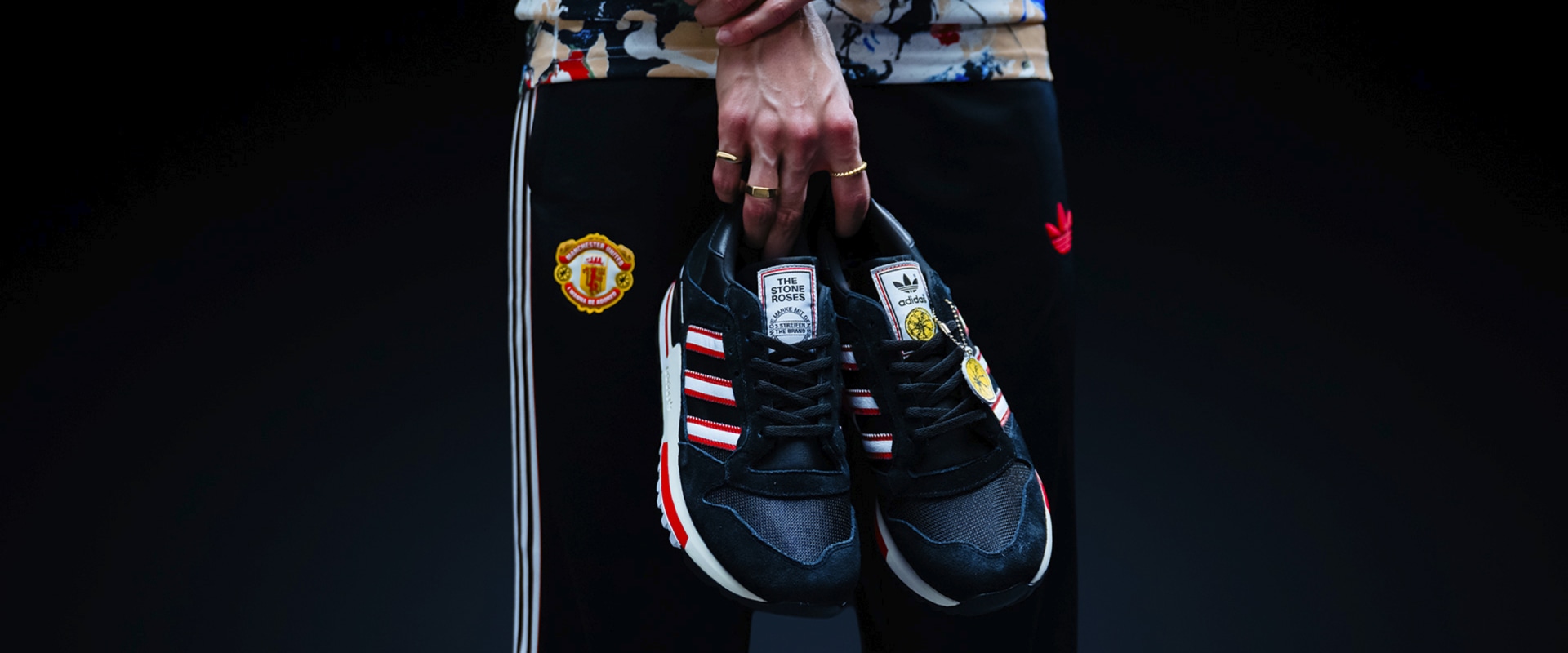 The image shows a pair of black and red athletic shoes with the Manchester United logo prominently displayed, set against a dark background.