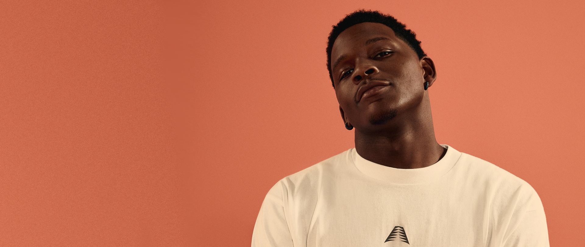 A young man with a serious expression stands against a vibrant orange background, wearing a plain white t-shirt.