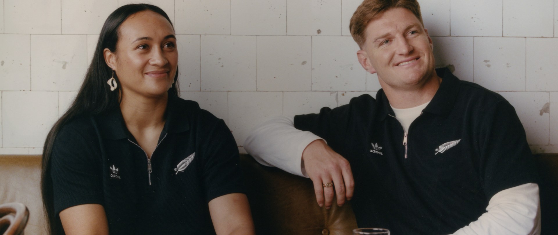 Two individuals, a woman and a man, are seated together wearing black hooded sweatshirts against a plain background.