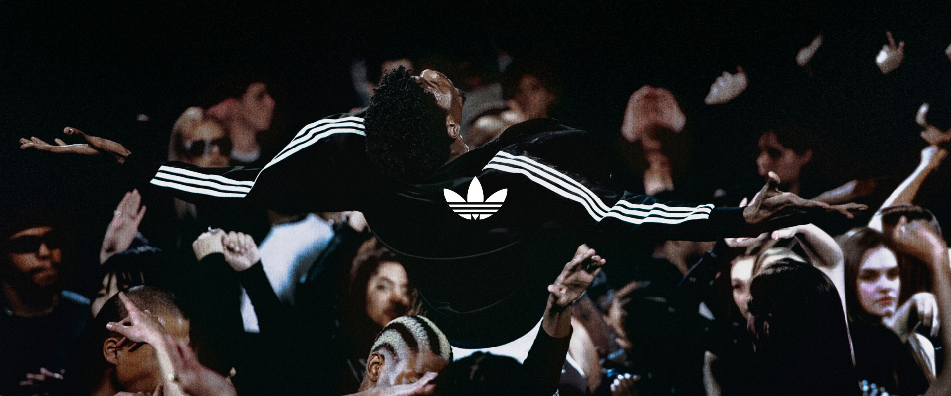 Person in a black Adidas Firebird jacket stands with arms outstretched in a crowded space. A white adidas trefoil logo is in the centre of the image.