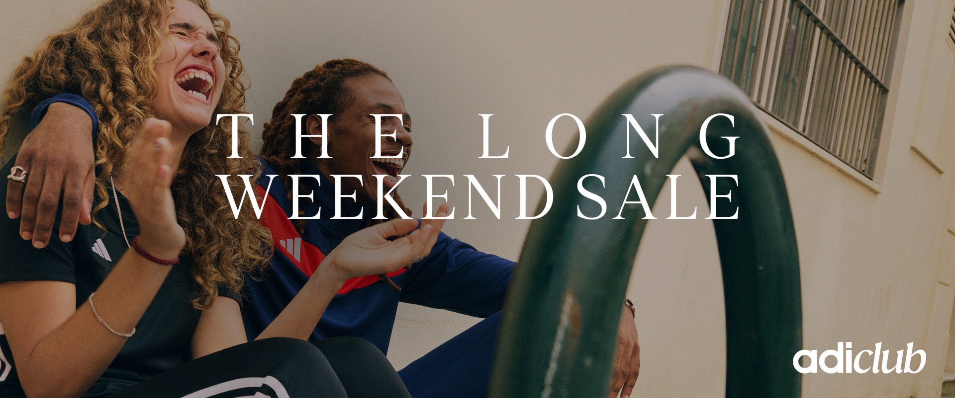 Two young women with curly hair, one laughing and the other smiling, sitting on a couch against a plain background, with the text "THE LONG WEEKEND SALE" overlaid on the image.