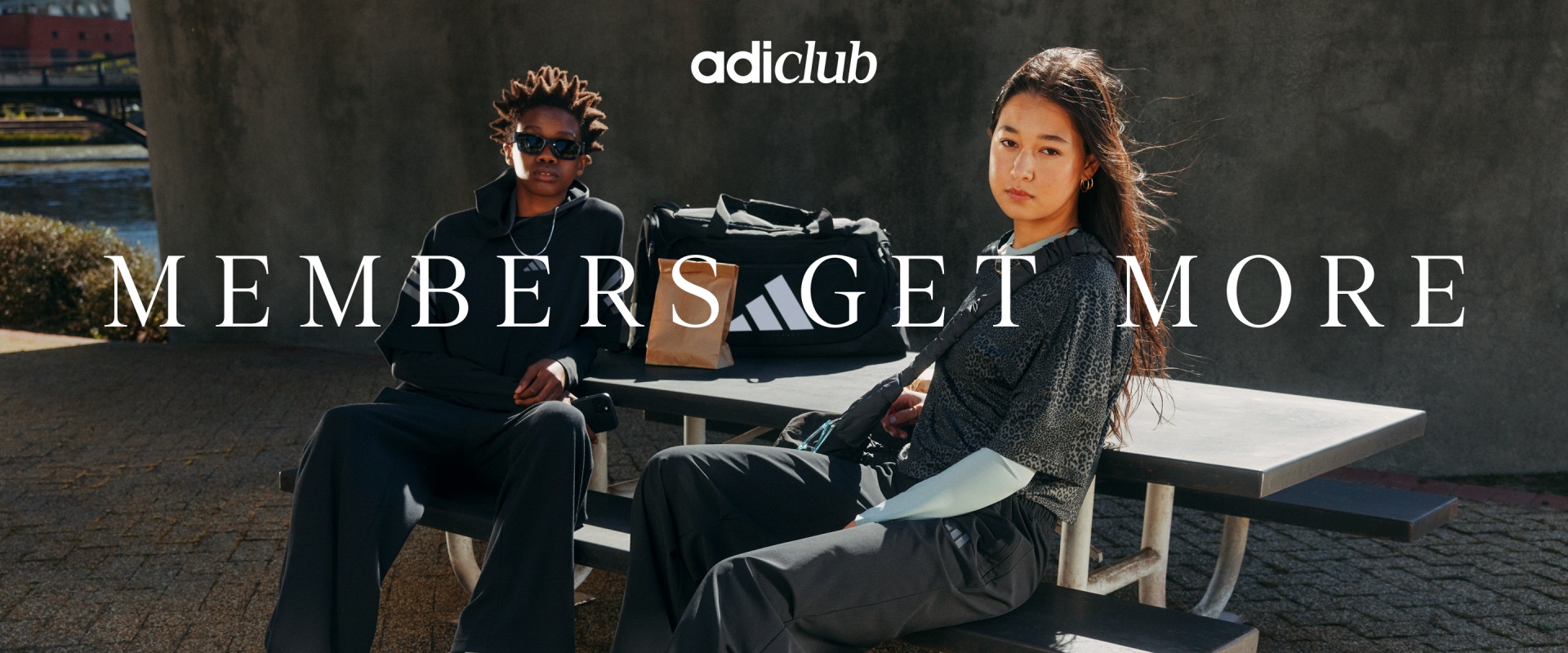 The image shows two individuals, a man and a woman, sitting at a table in front of a sign that reads "Members get more" and the Adclub logo.