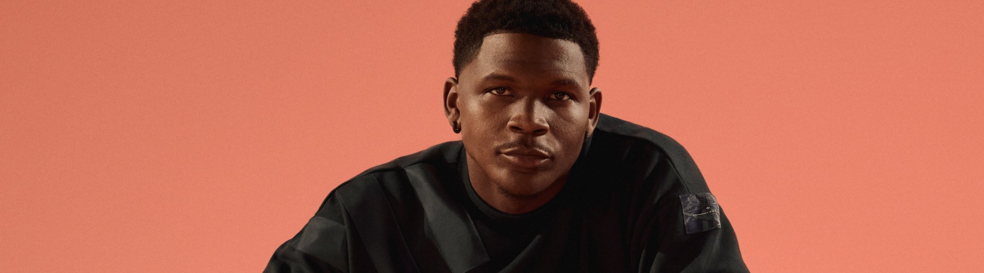 A young Black man with a serious expression, wearing a black shirt, against a vibrant coral-colored background.