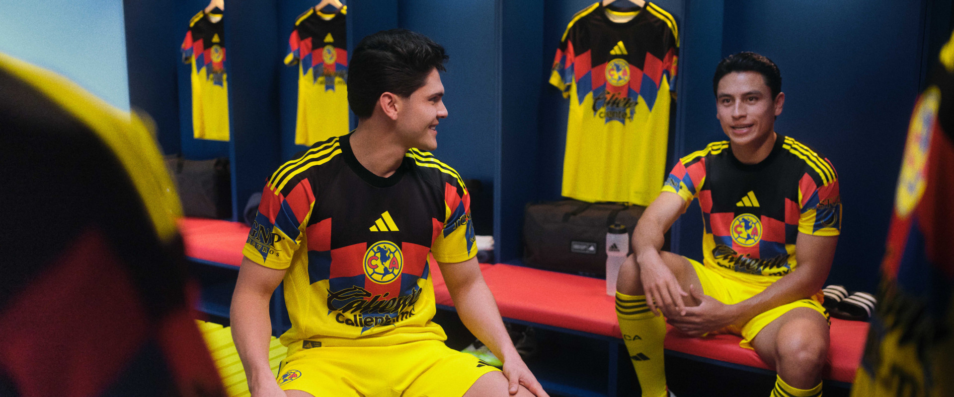 Two individuals wearing the national team's soccer jerseys are seated in what appears to be a locker room or training facility, with colorful team banners visible in the background.