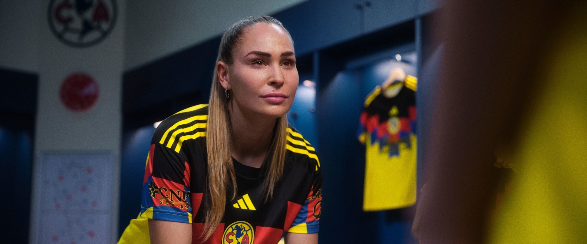 A young woman with long blonde hair wearing a colorful sports jersey stands in a room with sports-themed decor in the background.