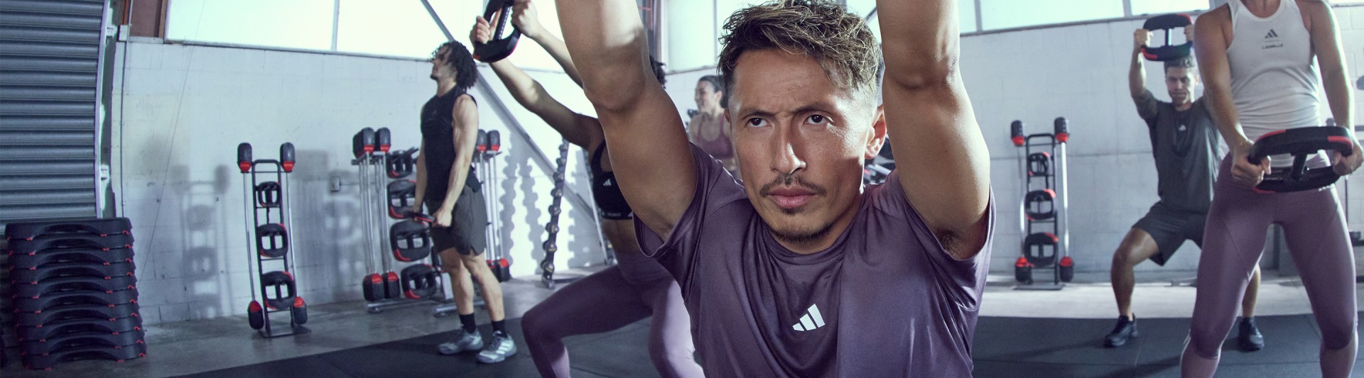 A man in a purple shirt is performing an exercise on a piece of gym equipment, with various other fitness equipment visible in the background.