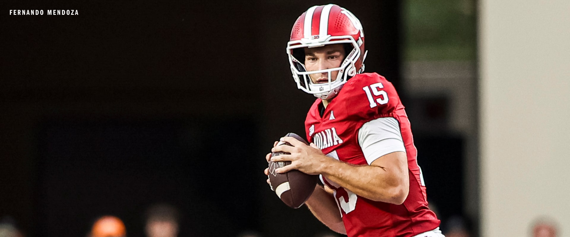 A football player in a red jersey and helmet stands on a field, holding a football.