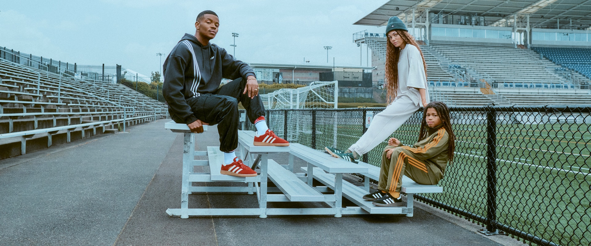 The image shows a group of three people, two women and one man, sitting on a set of bleachers in an outdoor sports stadium setting. The man is wearing a black jacket and the women are wearing casual clothing. The background includes a fenced-in field and stadium seating.