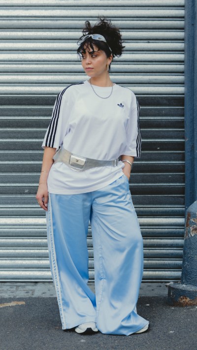 A person wearing a white t-shirt and blue wide-leg pants stands in front of a gray metal shutter backdrop.