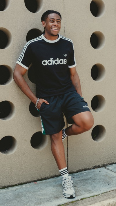 A smiling young man wearing an Adidas t-shirt and shorts, standing in front of a wall with circular patterns.