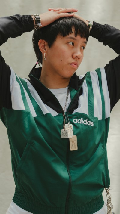 A young person wearing a green and white striped Adidas jacket, with their hands on their head, against a plain white background.