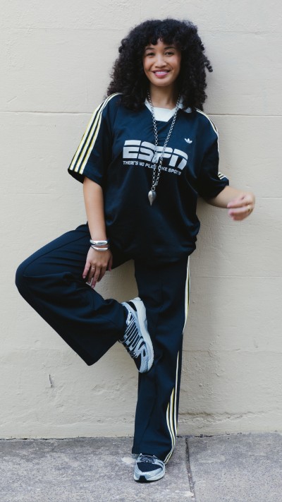 A young woman with curly hair wearing a navy blue Adidas tracksuit is standing against a white wall, smiling at the camera.