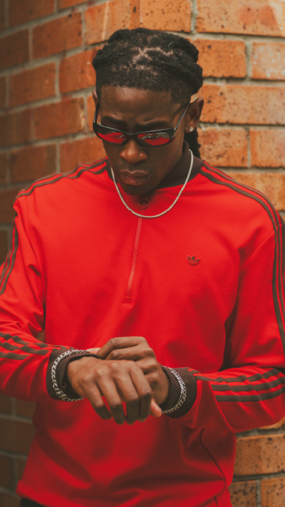 A young person wearing a bright red shirt and sunglasses stands in front of a brick wall.