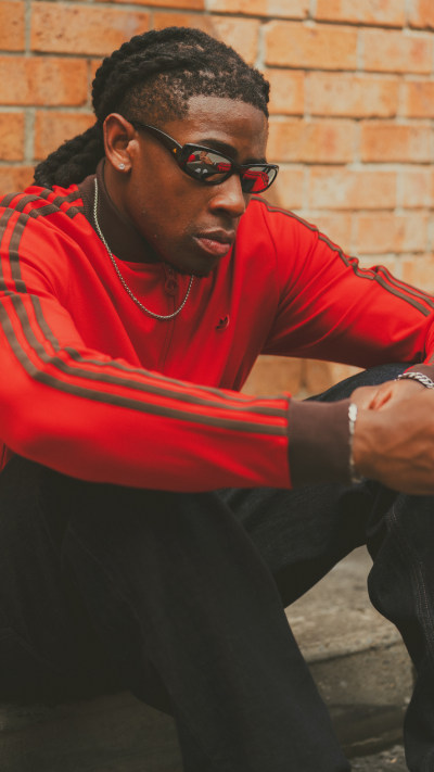 A young man with braided hair wearing a red shirt and glasses, sitting against a brick wall background.