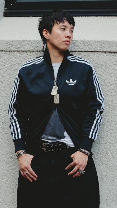 A young person wearing a black Adidas jacket with white stripes stands in front of a plain white wall.