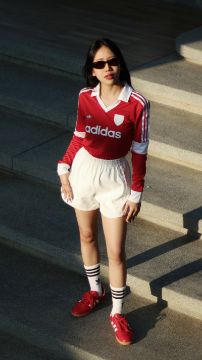 A young woman wearing a red Adidas jersey, white shorts, and red sneakers stands in front of a dark background.