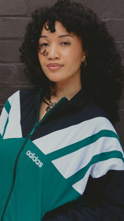 The image shows a young woman with curly dark hair wearing a green and white striped Adidas jacket, standing in front of a dark background.