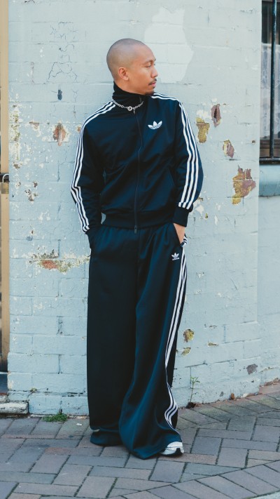 A person wearing a black and white tracksuit stands in front of a dilapidated brick wall.
