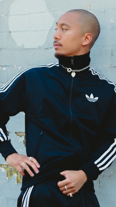 A person wearing a black Adidas tracksuit jacket stands in front of a white wall, with a serious expression on their face.