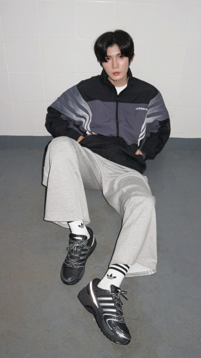 A person wearing a gray and black jacket and light-colored pants is seated on a gray floor, with a plain background behind them.