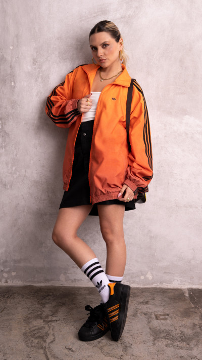 A young woman wearing an orange jacket and black shorts stands against a plain white wall, her expression serious as she poses for the camera.